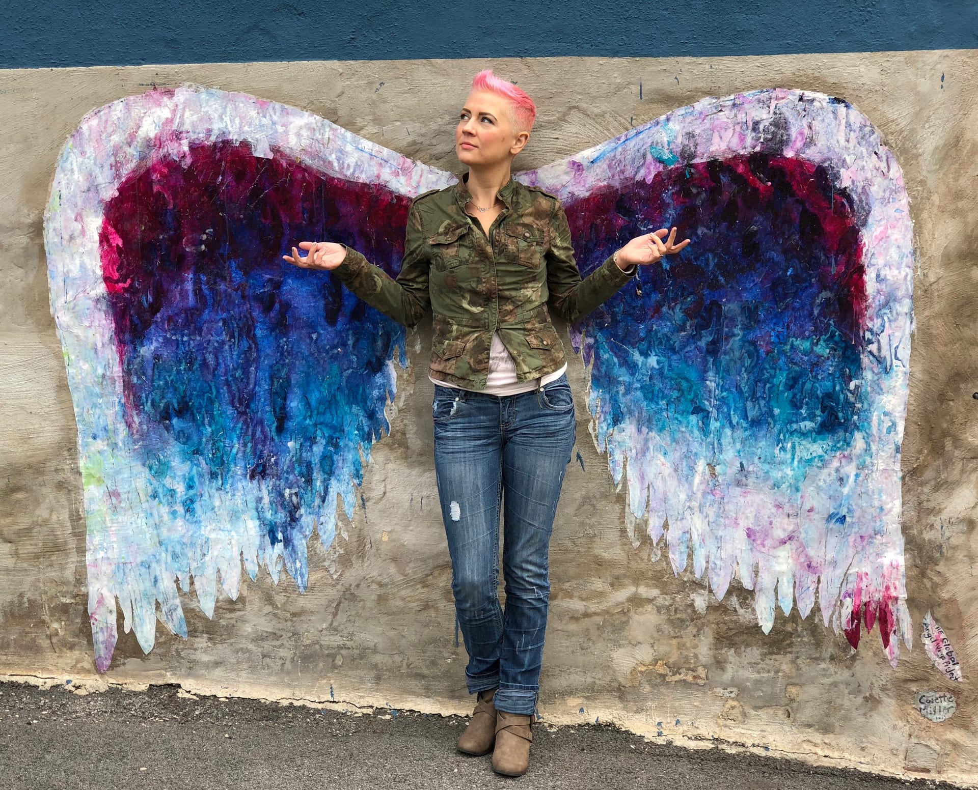 Blonnie standing against a wall mural of painted angel wings, arms open in front of them.