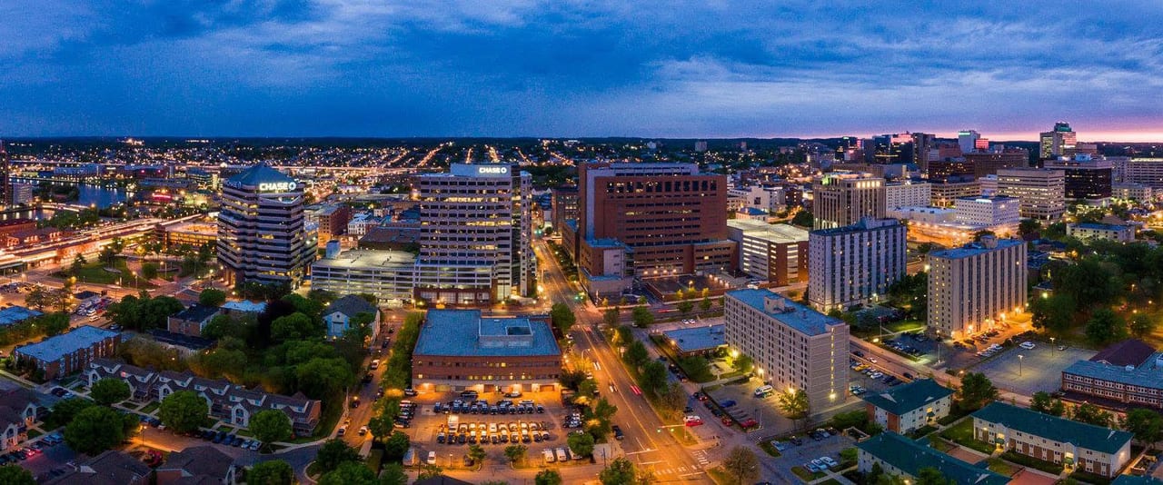 Aerial frame of a city at blue hour - lit office towers, streets running out toward the suburbs, a river along the left edge and the last of the sunset on the horizon.