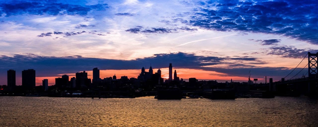 A city skyline in silhouette across the water at sunset, a suspension bridge lit at the right edge and the sky running from orange to deep blue.